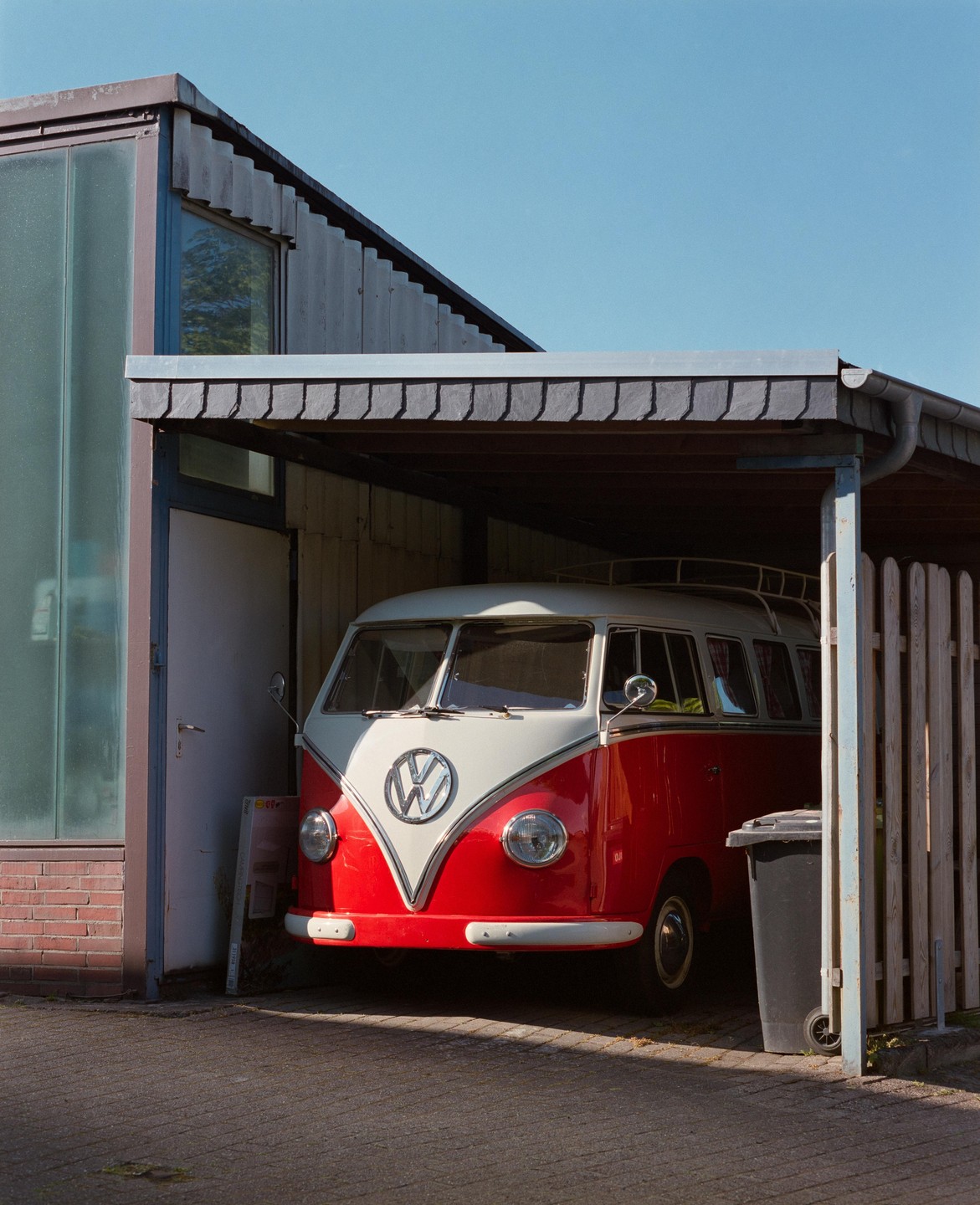 A red and white Volkswagen van is parked under a carport next to a building with a glass facade.