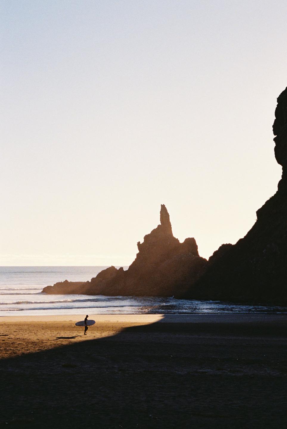 A surfer carrying a surfboard walks along a beach with large rock formations at sunset.