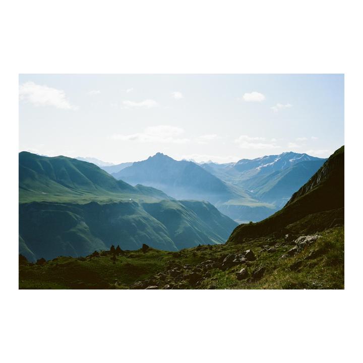 A vast mountain landscape shows green slopes in the foreground and hazy, snow-capped peaks in the distance under a bright sky.