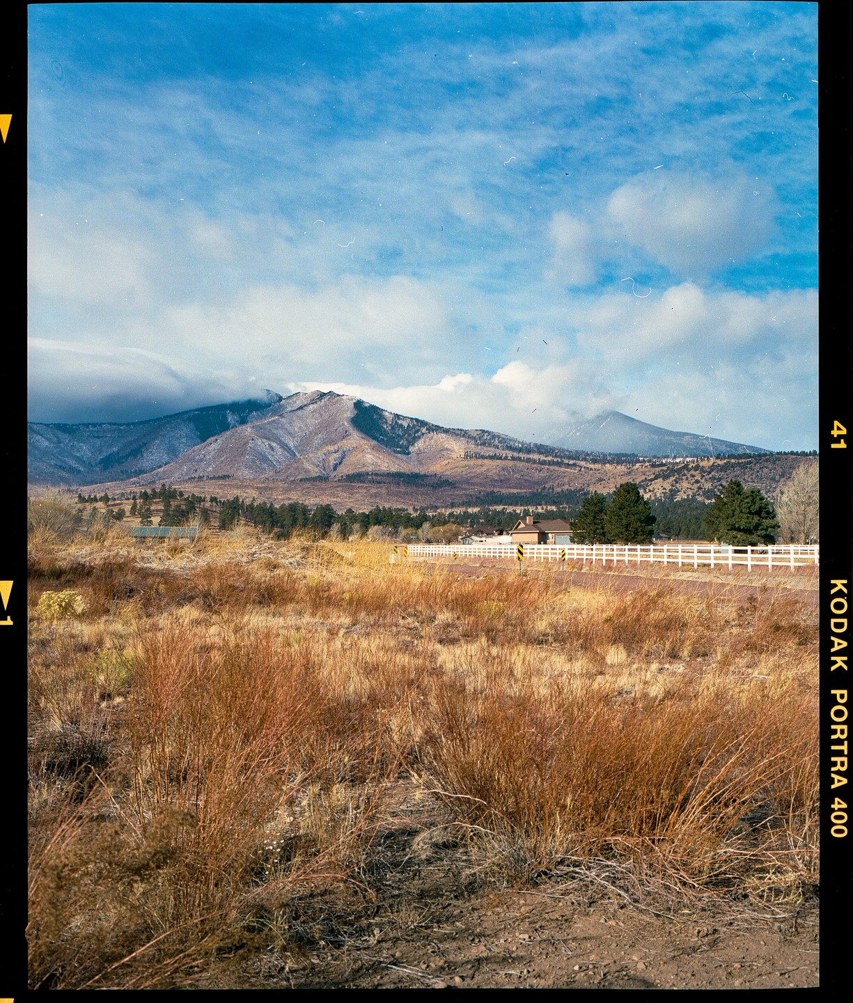 A landscape shows a mountain range with snow-capped peaks under a blue sky with clouds, with a field of dry grass and a white fence in the foreground.