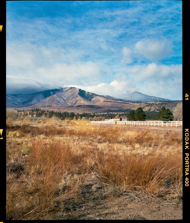 A landscape shows a mountain range with snow-capped peaks under a blue sky with clouds, with a field of dry grass and a white fence in the foreground.