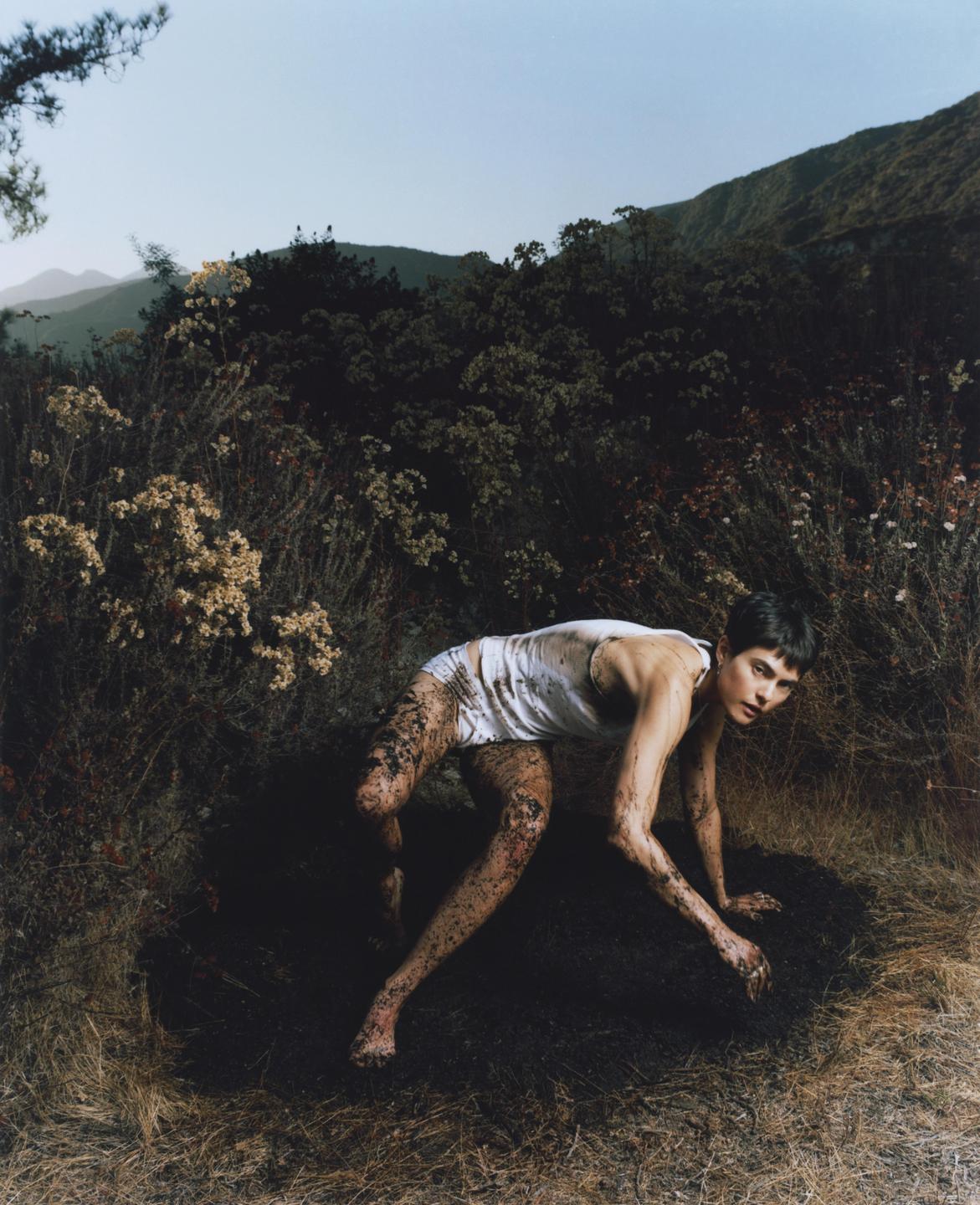 A woman covered in mud crawls on the ground in a field with mountains in the background.
