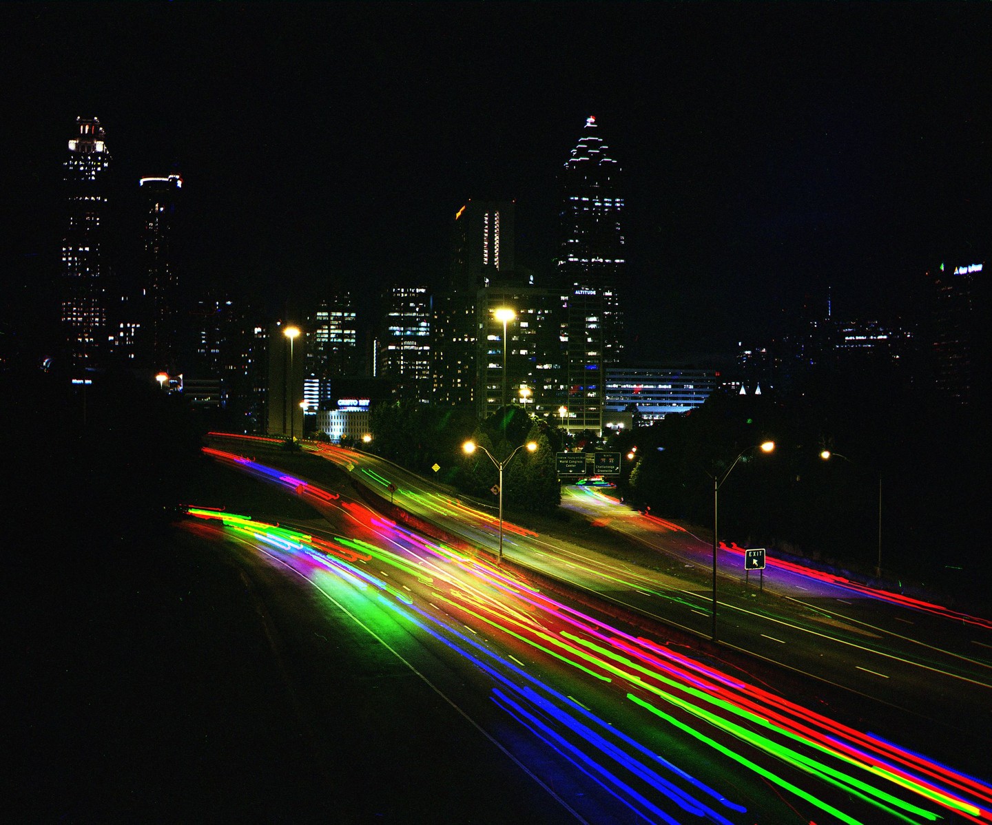 Light trails from vehicles on a highway at night lead towards a city skyline.