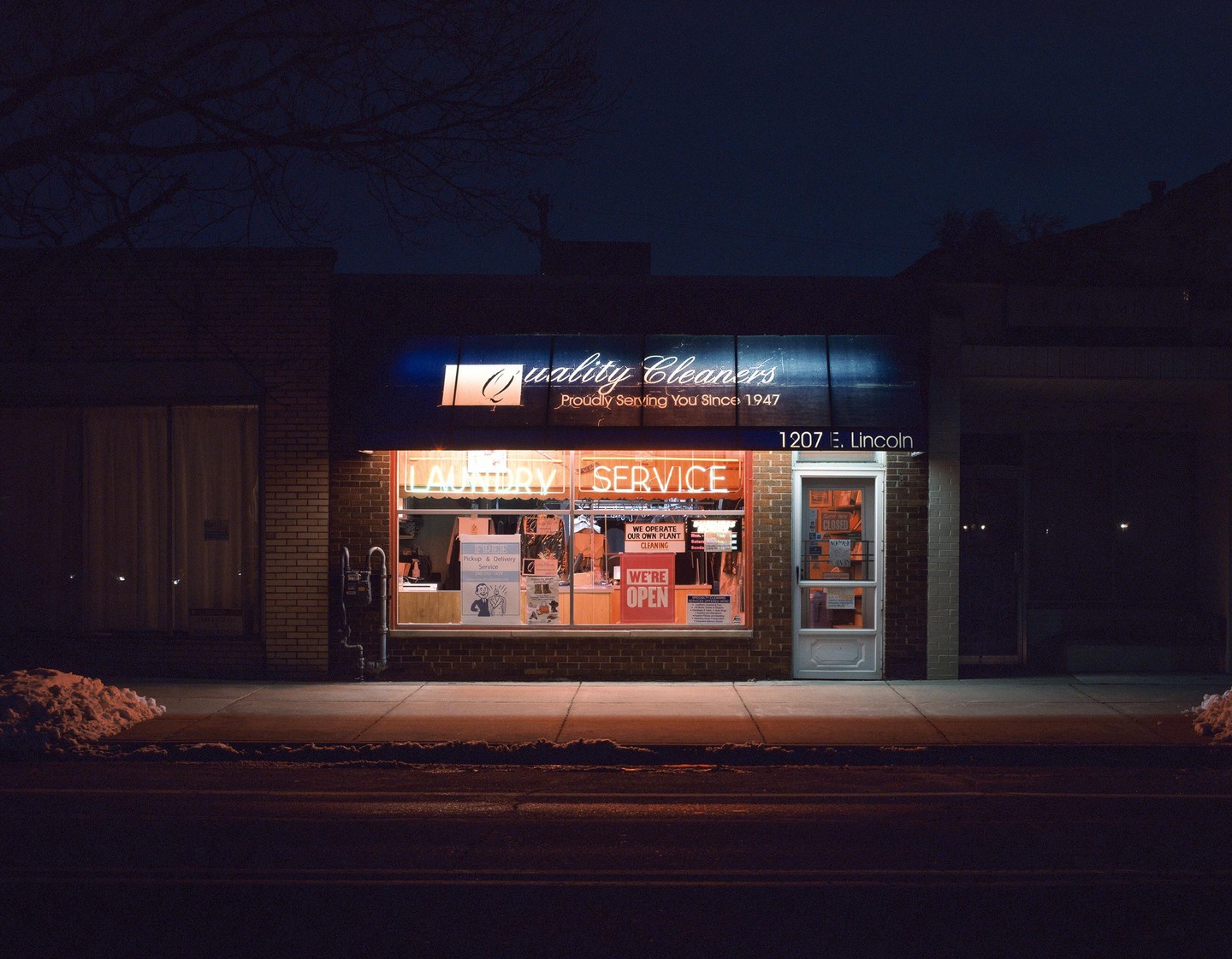 A brightly lit laundry service storefront glows at night, with a small pile of snow on the sidewalk.