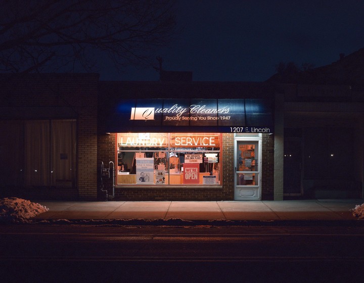 A brightly lit laundry service storefront glows at night, with a small pile of snow on the sidewalk.