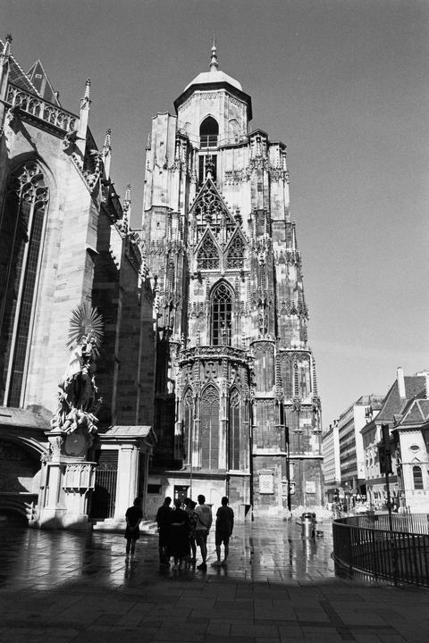 A group of people stand on a wet street in front of a large, ornate gothic cathedral.
