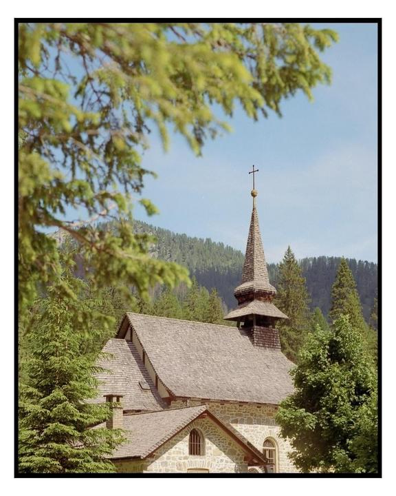 A stone church with a tall steeple and cross is nestled among green trees with mountains in the background.