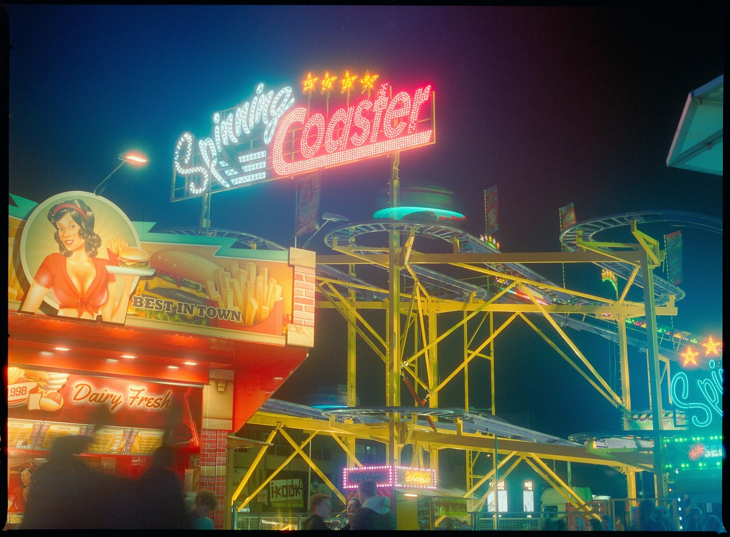 A neon sign for a spinning coaster ride is illuminated at night next to a food stall with a retro-style illustration of a woman holding a burger and fries.