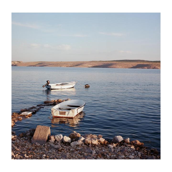 Two small boats float on the water near a rocky shore with a distant landmass under a clear sky.
