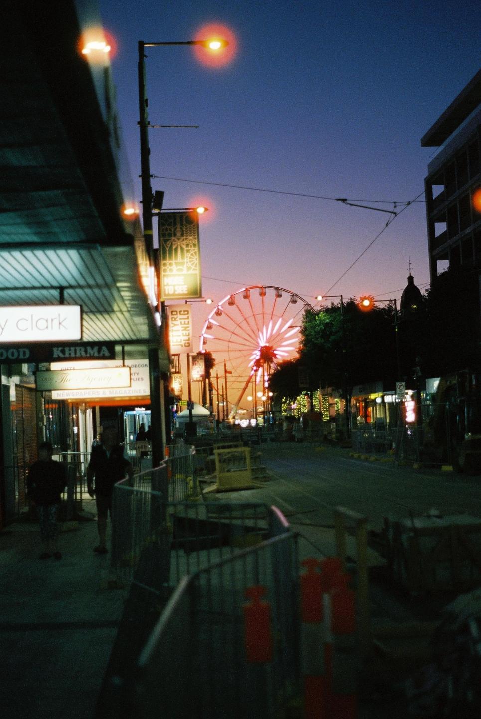 A street at dusk is illuminated by a ferris wheel and streetlights.
