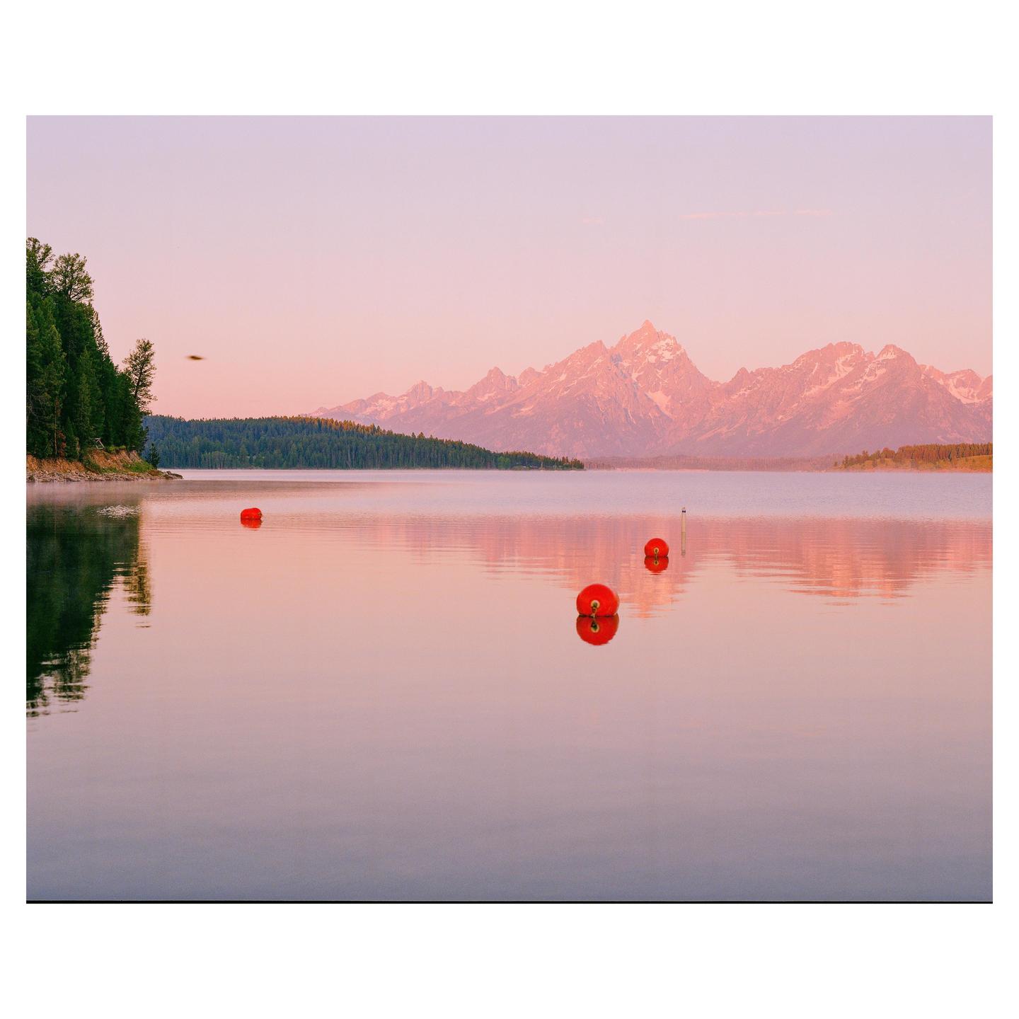 Three red buoys float on a calm lake with mountains and trees in the background under a pink sky.
