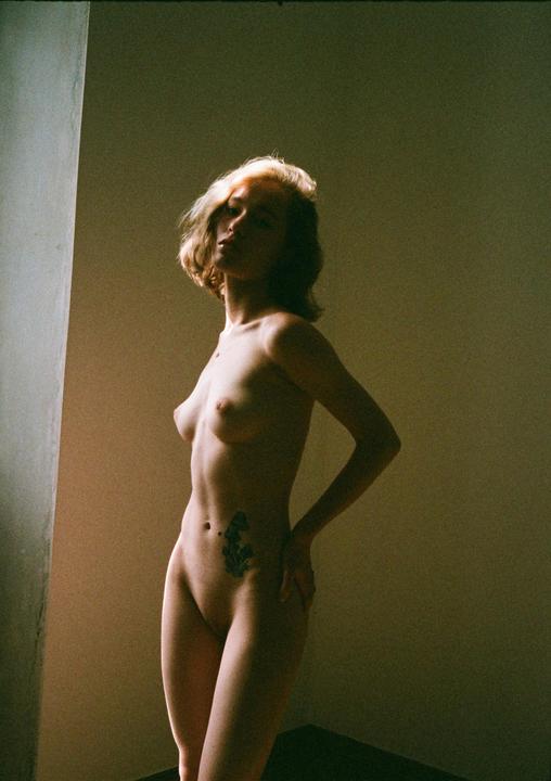 A woman with short hair and a tattoo on her hip stands in a room with light falling on her face and body.