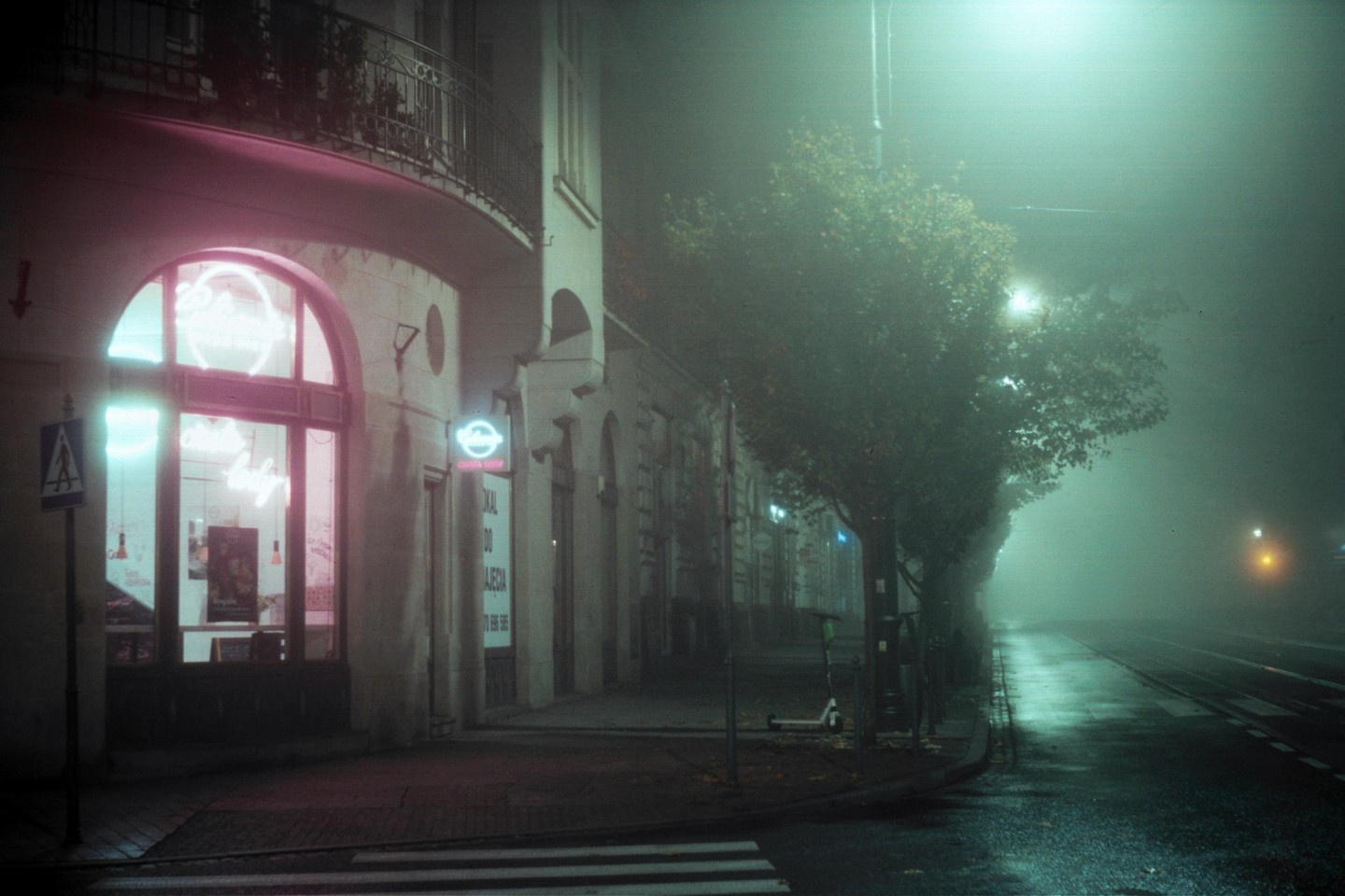 A foggy street at night features a building with neon signs and a tree-lined sidewalk next to a wet road.