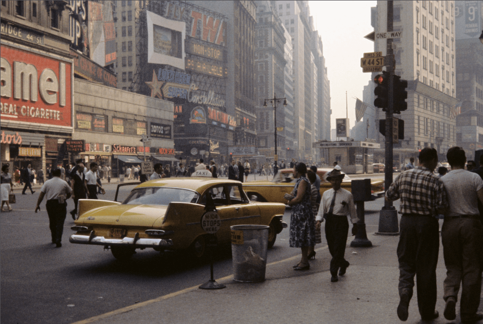 A busy street scene in New York City shows pedestrians, yellow taxis, and numerous billboards and signs on tall buildings.