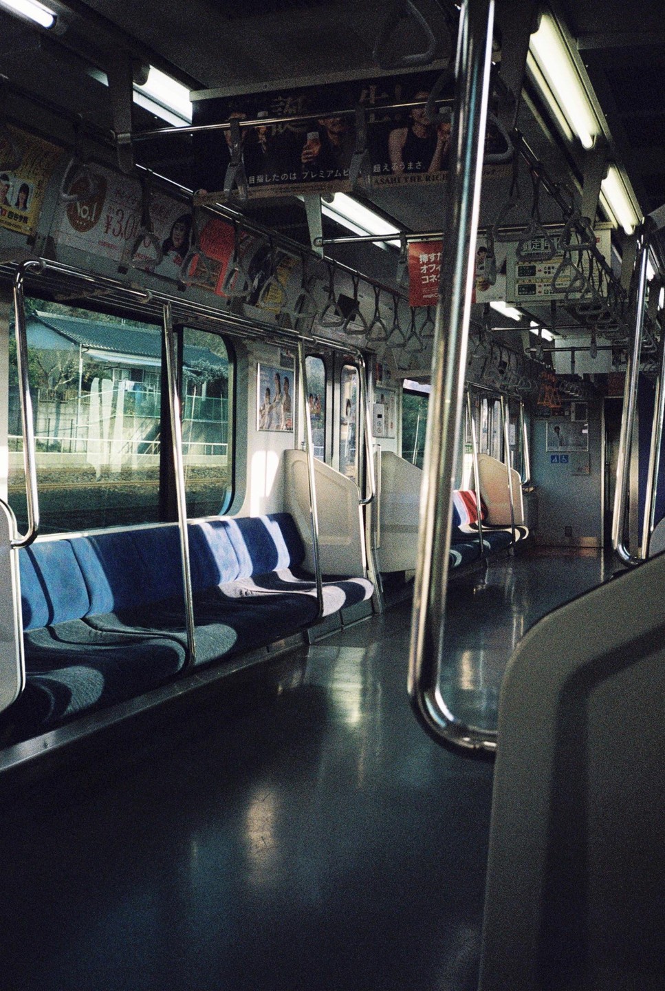 The interior of an empty train car with blue seats and advertisements on the ceiling is illuminated by sunlight streaming through the windows.