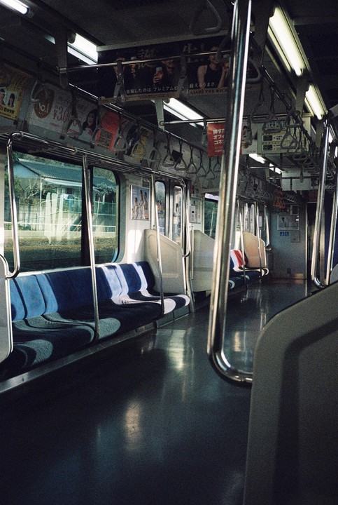 The interior of an empty train car with blue seats and advertisements on the ceiling is illuminated by sunlight streaming through the windows.