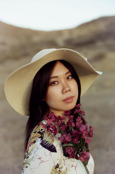 A woman wearing a wide-brimmed hat and a floral shirt holds a bouquet of purple flowers.