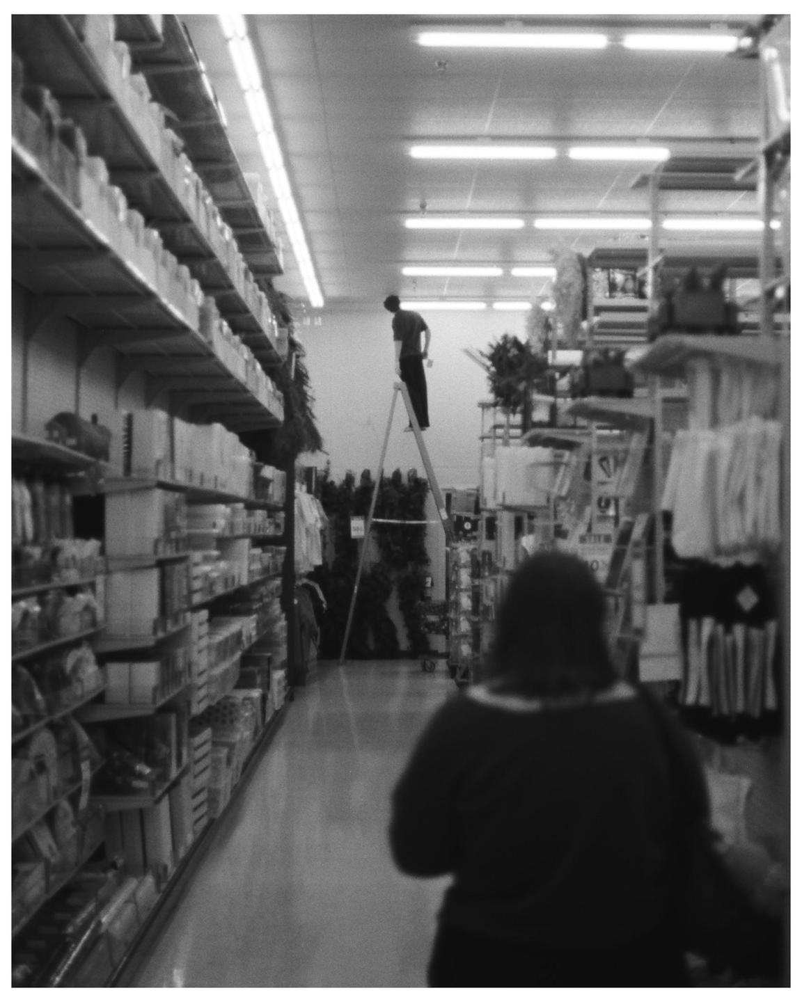 A man stands on a ladder in an aisle of a store while a woman walks away in the foreground.