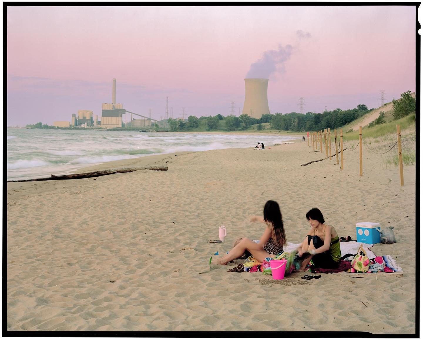 Two women sit on a sandy beach with a power plant and nuclear cooling tower in the background.