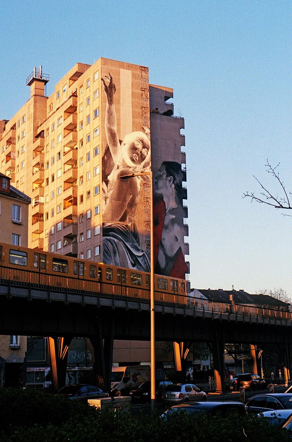 A yellow train passes on an elevated track in front of a building with a large mural.