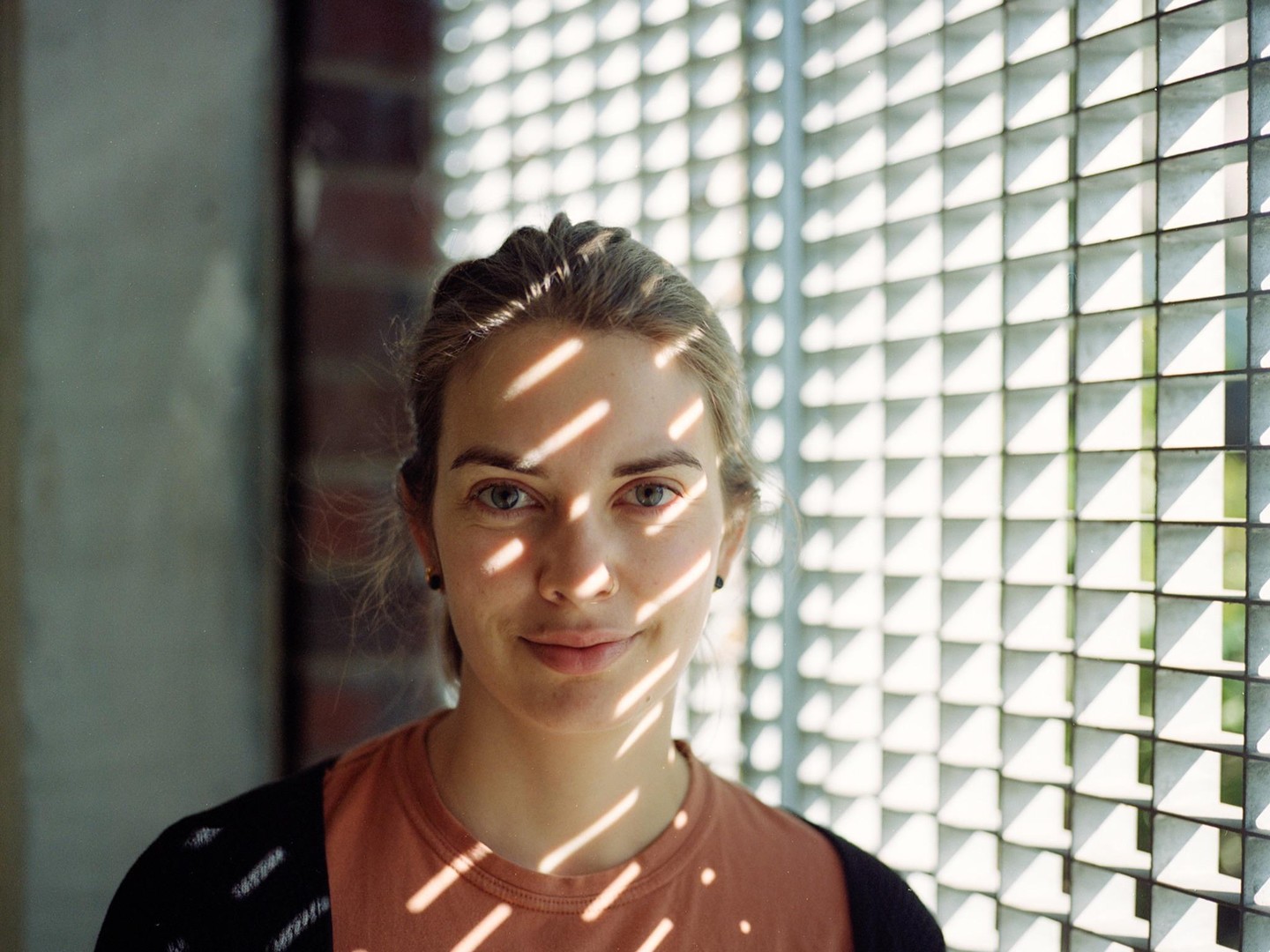 A woman with light hair and a nose ring smiles at the camera, with shadows from a grid pattern falling across her face and body.