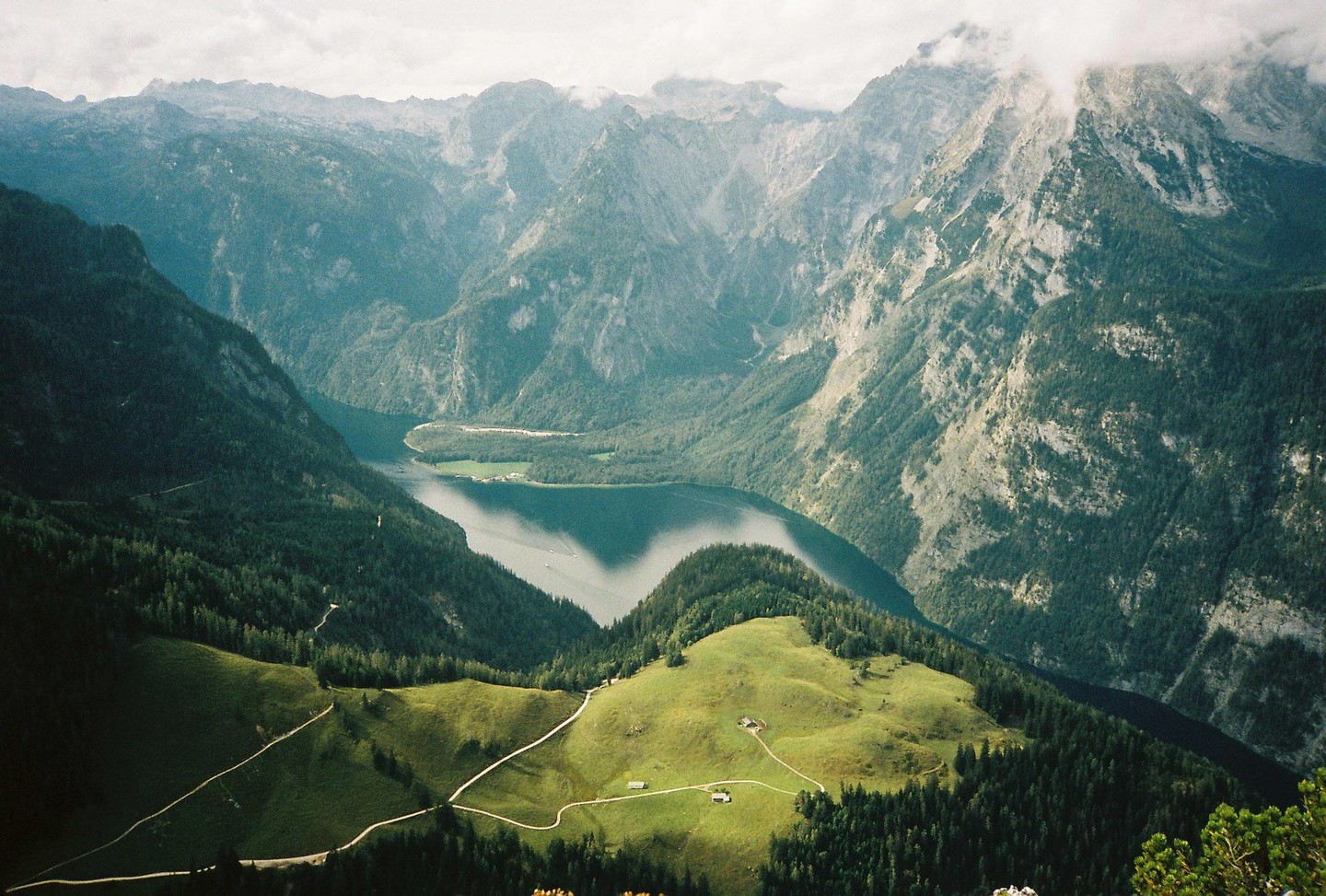 A winding road leads to a small house on a green hill overlooking a lake surrounded by mountains.