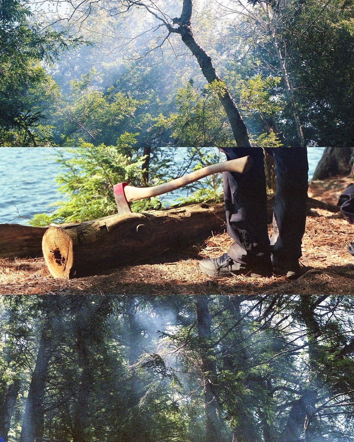 An axe is stuck in a log next to a person's legs, with a body of water and forest in the background.