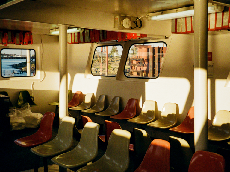 The interior of a ferry boat is filled with rows of empty seats in red and yellow, with sunlight streaming through the windows.