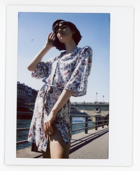A woman in a floral dress stands by a river with a bridge in the background.