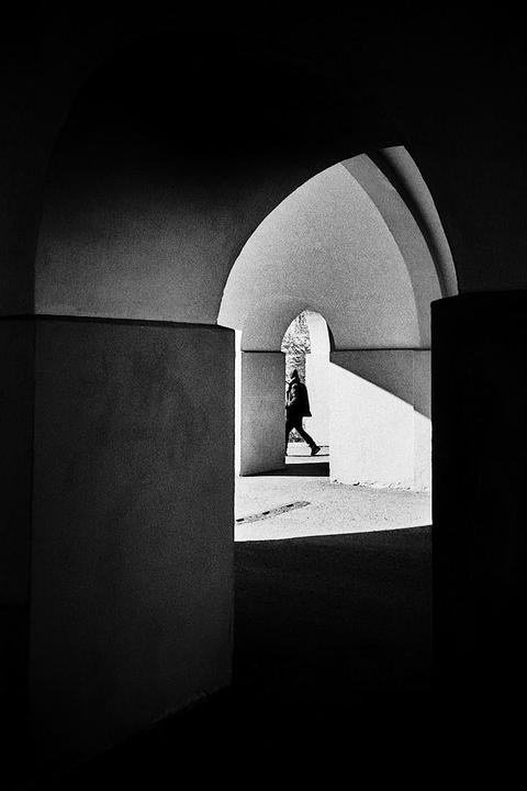 A person walks through a series of arches with strong shadows and highlights.