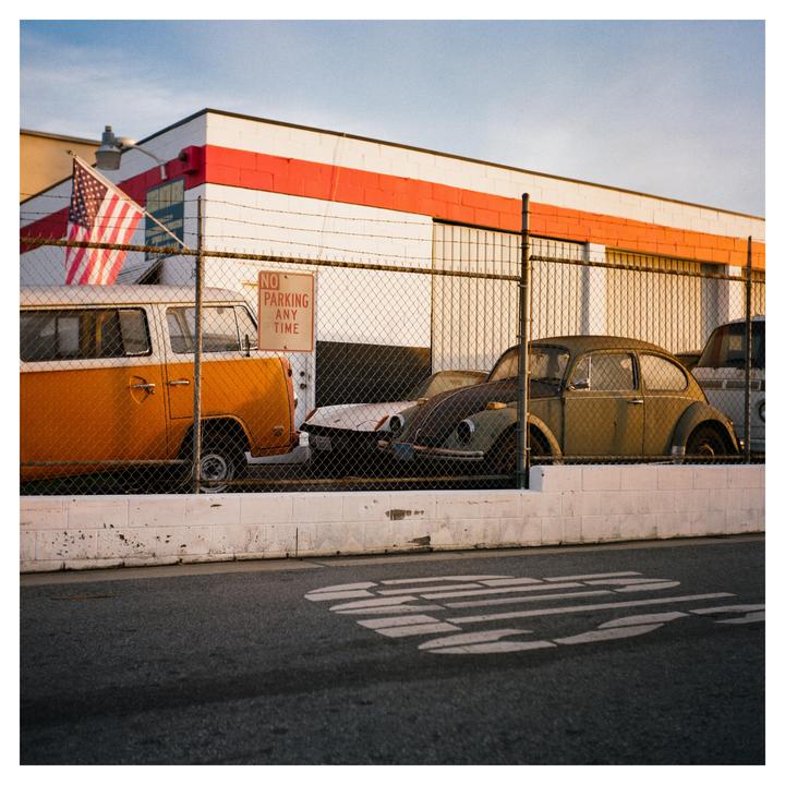 An orange van and a green beetle car are parked behind a chain-link fence with an American flag and a no parking sign.