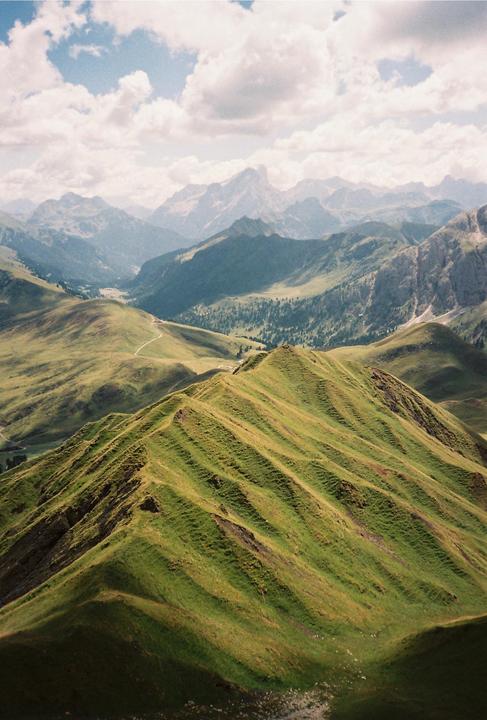 A winding path leads through a valley surrounded by green, rolling hills and distant mountains under a cloudy sky.