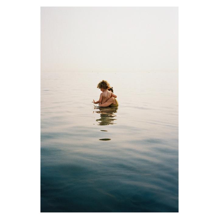 A child with curly hair sits in calm water, facing away from the viewer.