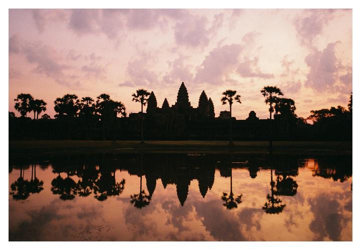 A silhouette of Angkor Wat and palm trees is reflected in water under a cloudy, colorful sky.