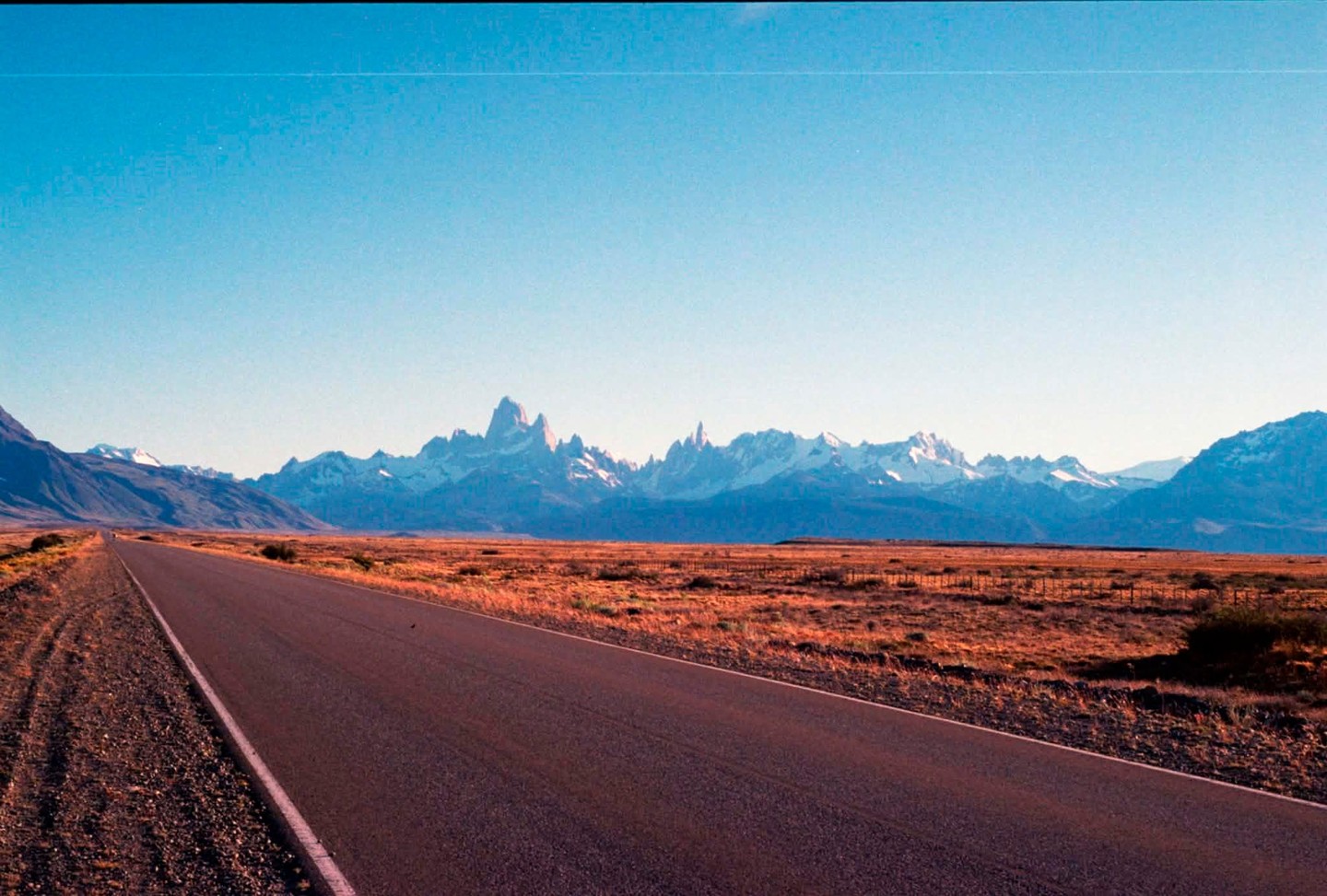 A long, empty road stretches towards a distant range of snow-capped mountains under a clear sky.