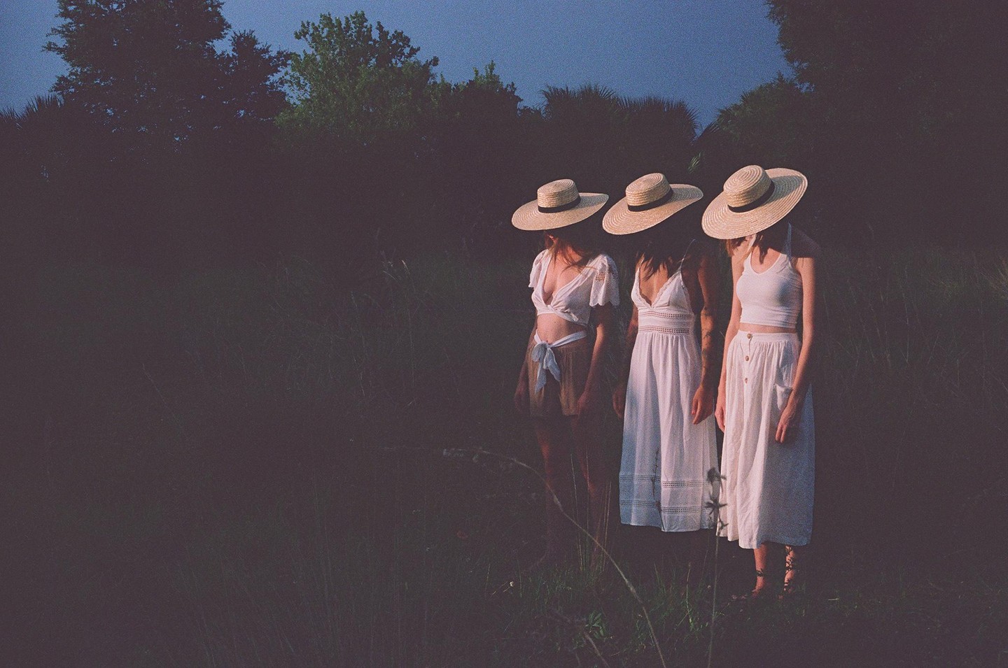 Three women in white dresses and straw hats stand in a field at dusk.
