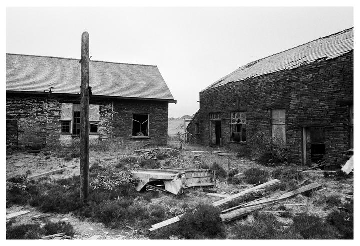 Two derelict stone buildings with broken windows stand in a field of overgrown grass and scattered debris.