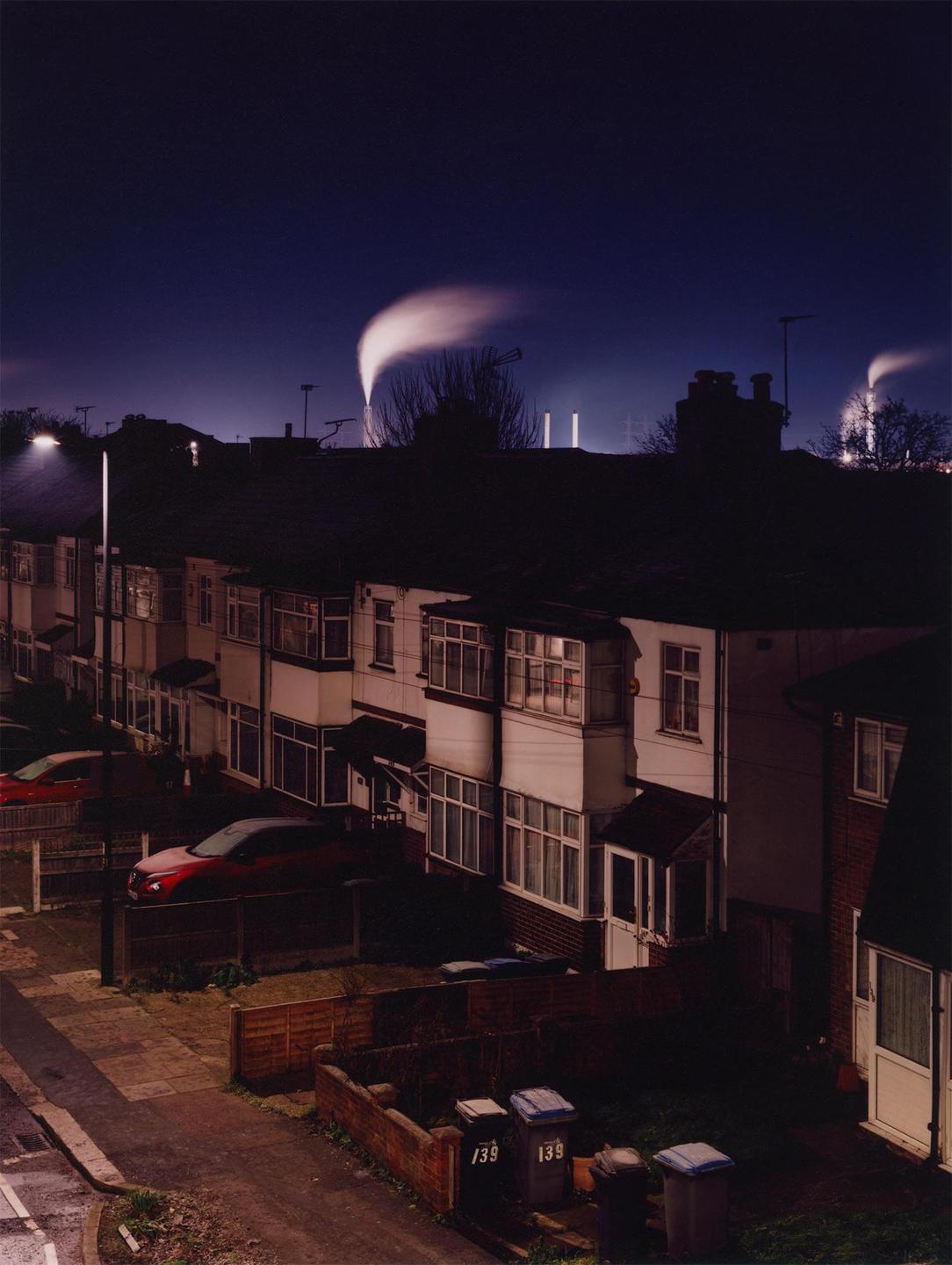 A row of houses with bay windows and parked cars is visible at night, with industrial smoke plumes rising in the distance.