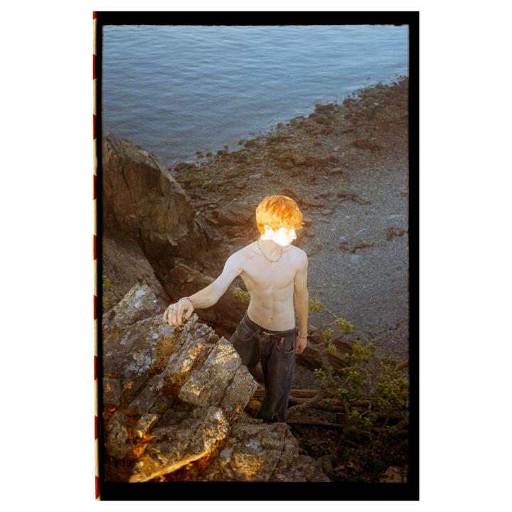 A shirtless man with red hair stands on a rocky cliff overlooking the ocean.