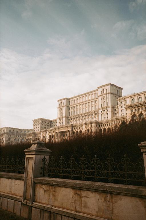 A large, ornate building with many windows and arches stands behind a decorative metal fence and stone wall.