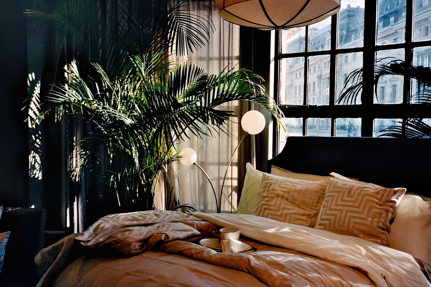 Sunlight streams into a bedroom with a large plant, a bed with pillows, and a window looking out onto city buildings.