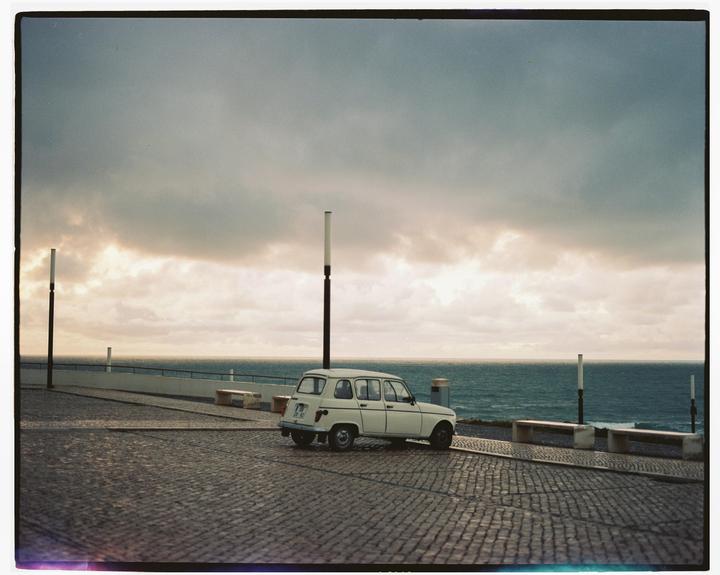 A white car is parked on a cobblestone street overlooking the ocean under a cloudy sky.