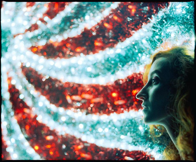 A woman's profile is visible on the right side of the frame, with a blurred background of red and blue tinsel.