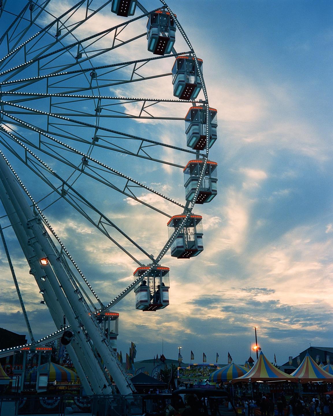 A ferris wheel with illuminated gondolas stands against a cloudy sky at dusk, with carnival tents and people visible below.