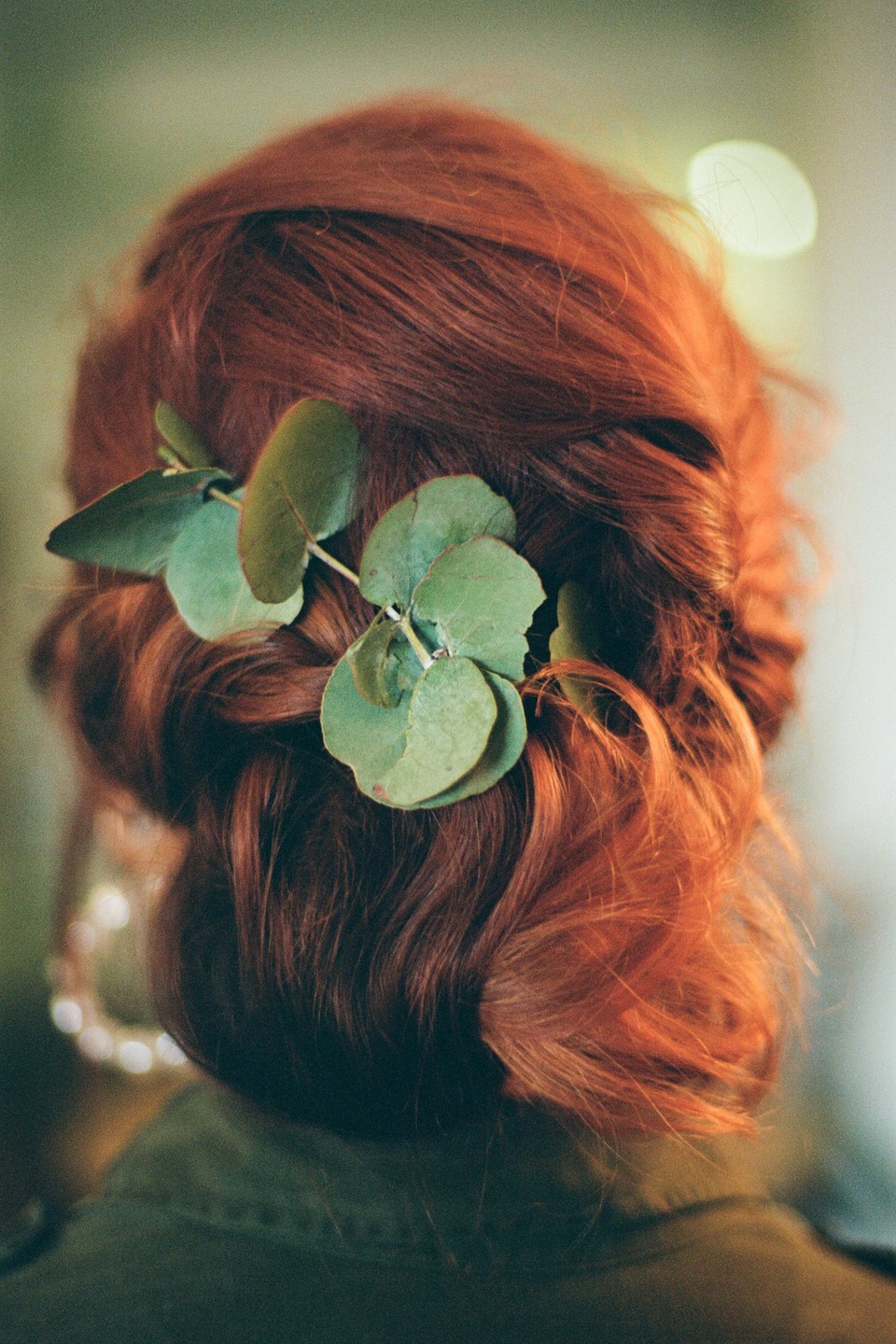A woman's red hair is styled in an updo with a sprig of eucalyptus leaves tucked into it.
