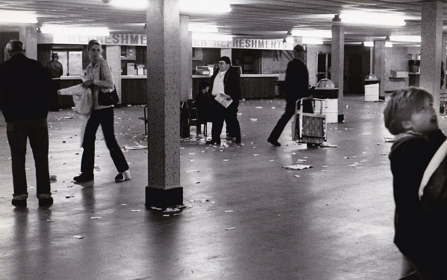 People walk across a dirty floor in a large room with refreshment stands.