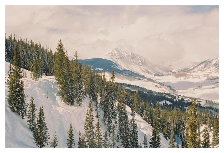 A snow-covered mountain slope with pine trees descends into a valley with distant mountains under a cloudy sky.