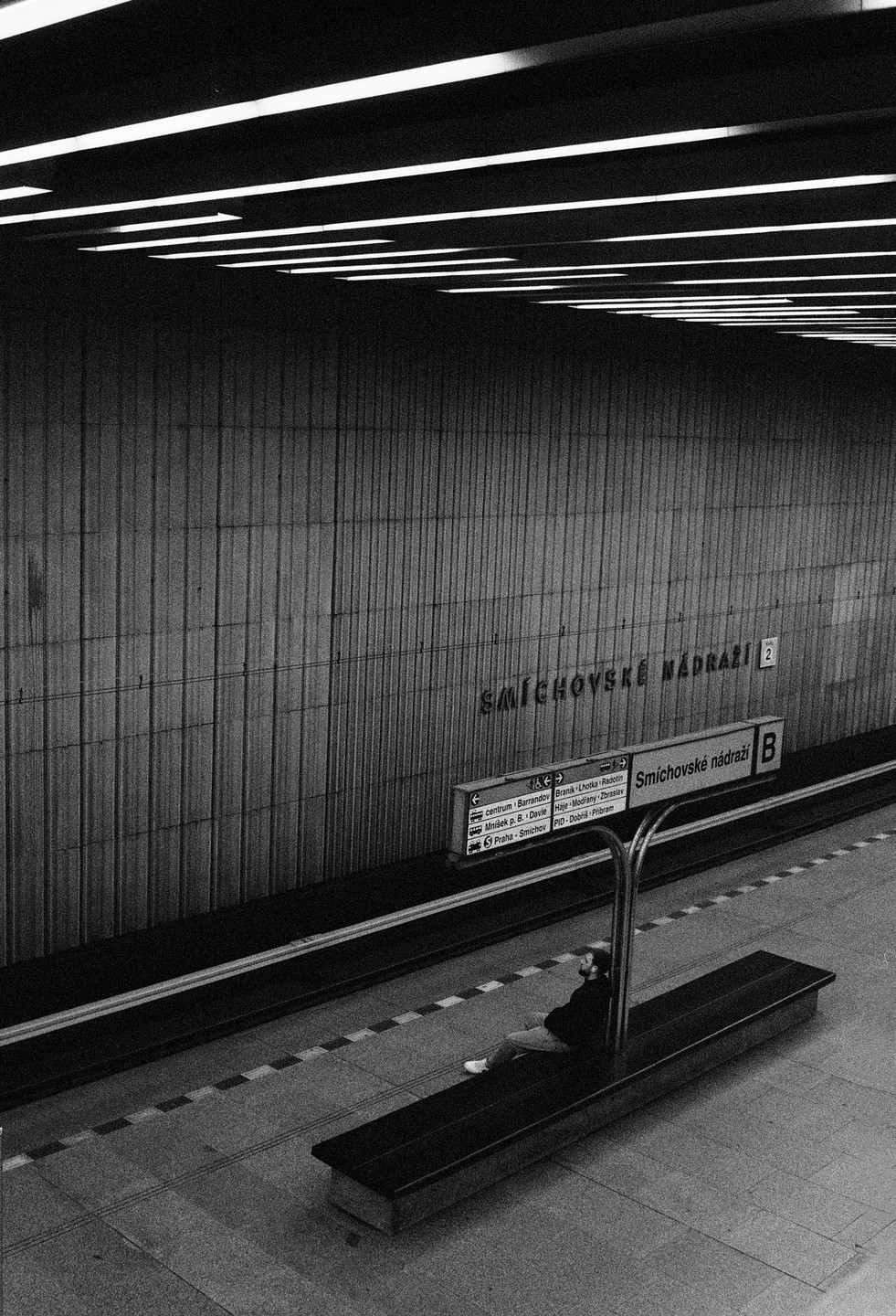 A man sits on a bench in a subway station with a sign for Smichovske Nadrazi.