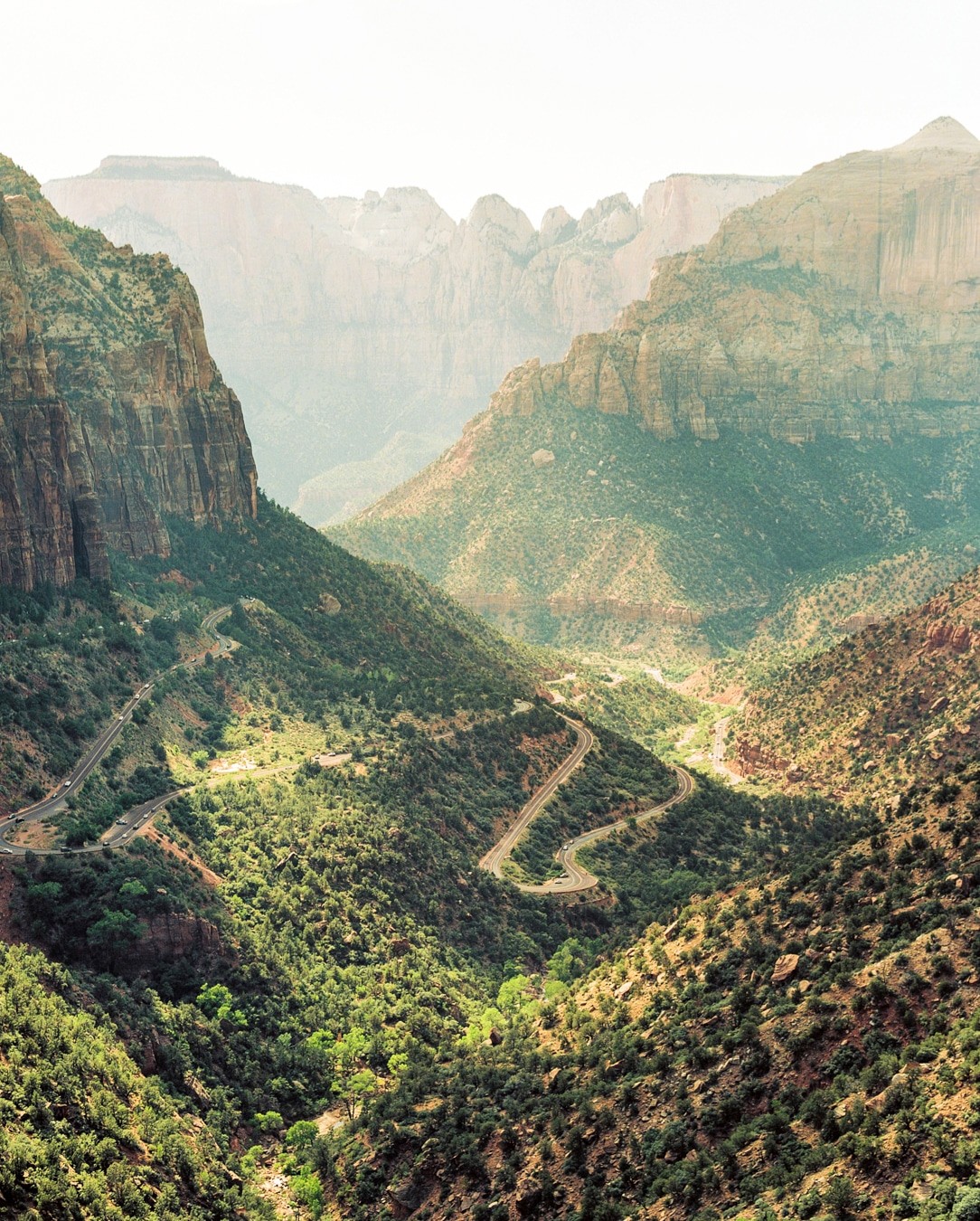A winding road descends through a valley surrounded by mountains and green foliage.