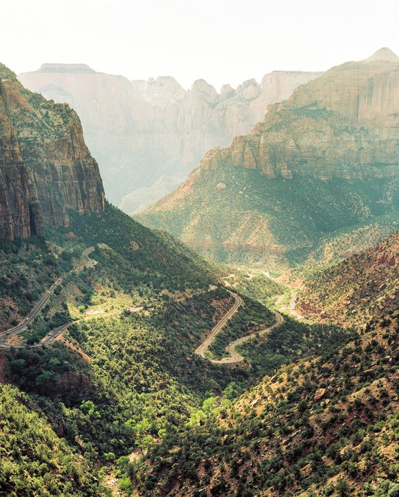 A winding road descends through a valley surrounded by mountains and green foliage.